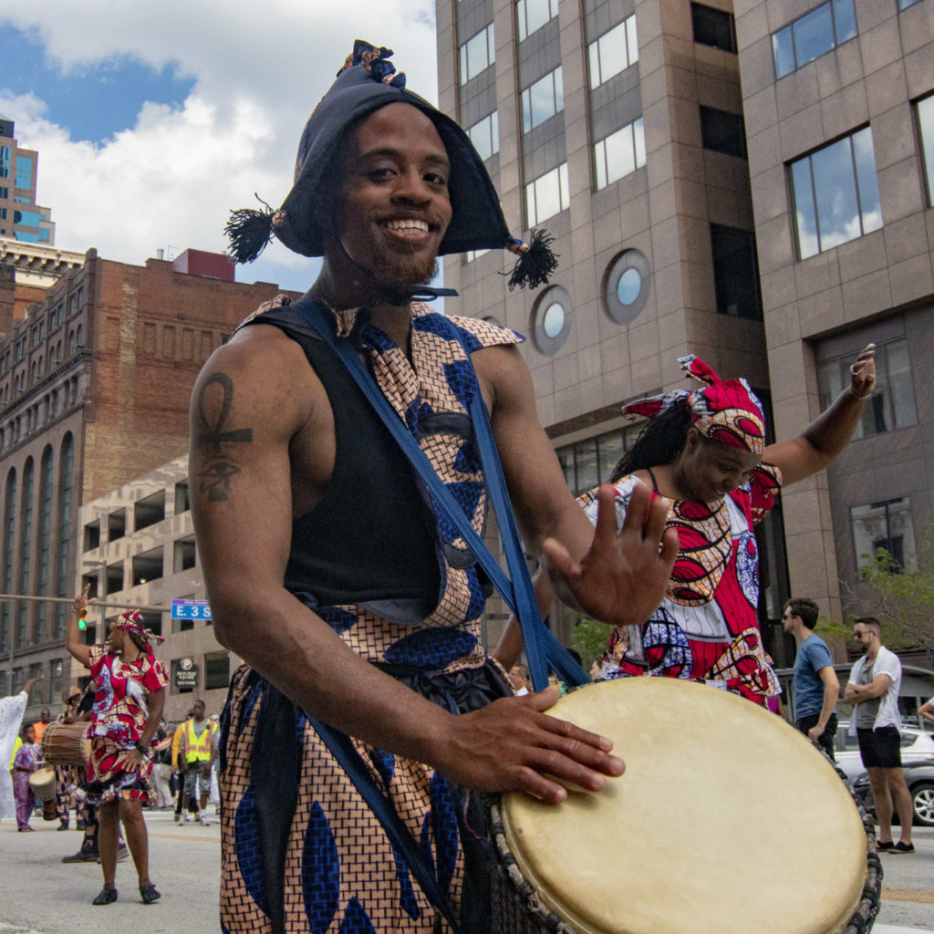 Performer smiling while playing a drum during a BorderLight Theatre Festival street parade in downtown Cleveland.